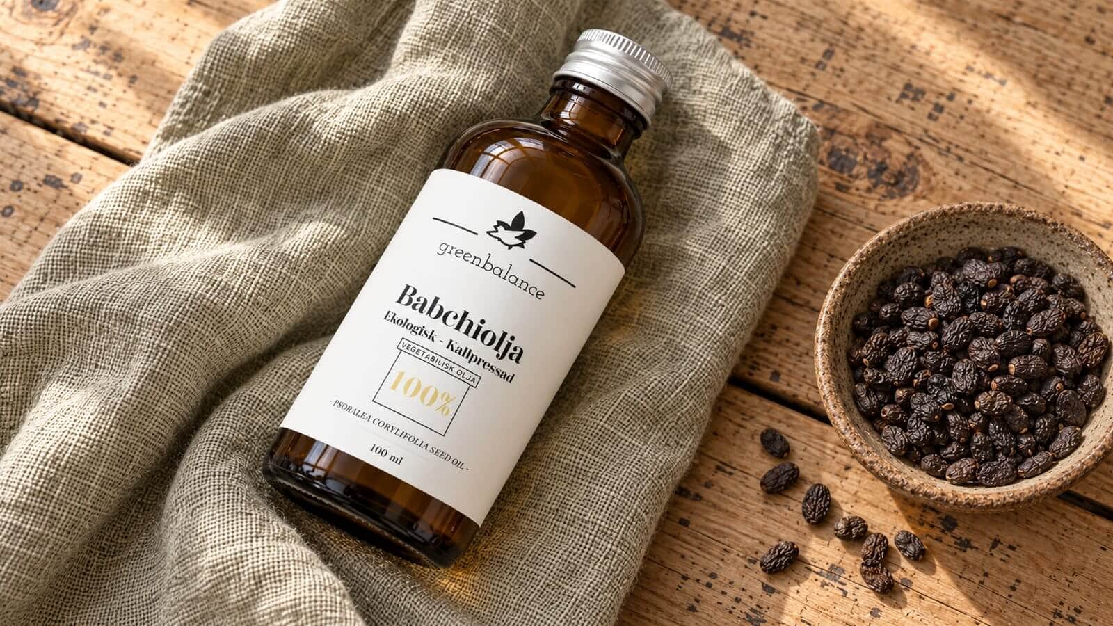 babchiolja bakuchiol produkt ravaror Flaska med babchiolja liggande på linne och trä, med mörka babchifrön i en skål.