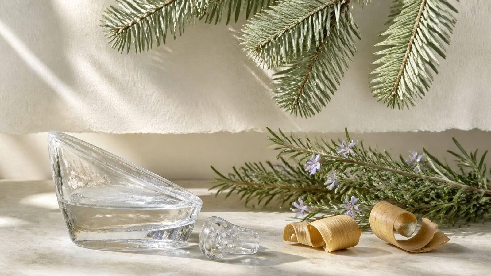 Silvergröna granbarr, blommande rosmarin, träspån och klar vätska i glas.