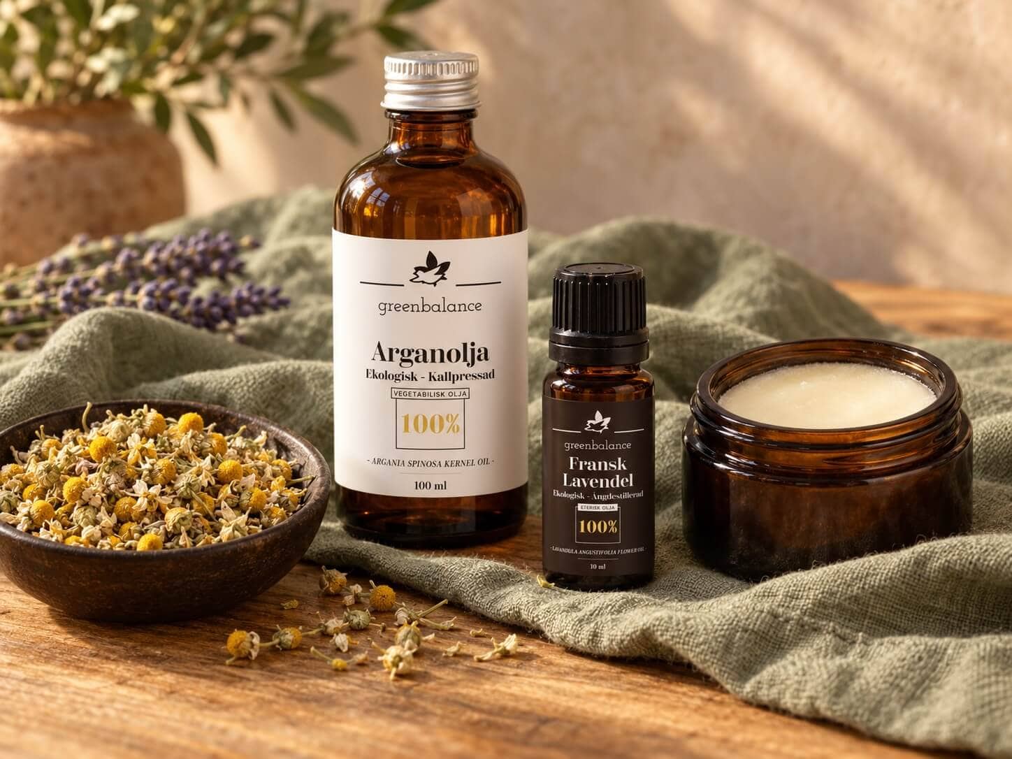 Hero – råvaror Greenbalances arganolja och franska lavendelolja med kamomill och en öppen burk sheasmör på grönt linne och trä