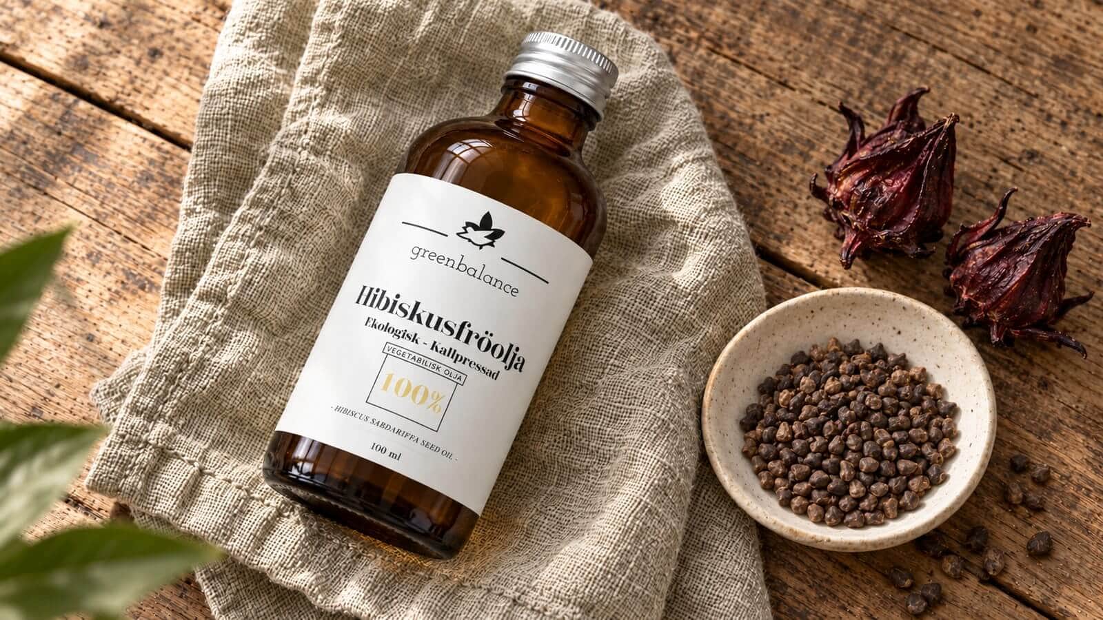 hibiskusfroolja produkt ravaror Flaska med hibiskusfröolja liggande på linne och trä, med hibiskusfrön och torkade blomfoder.
