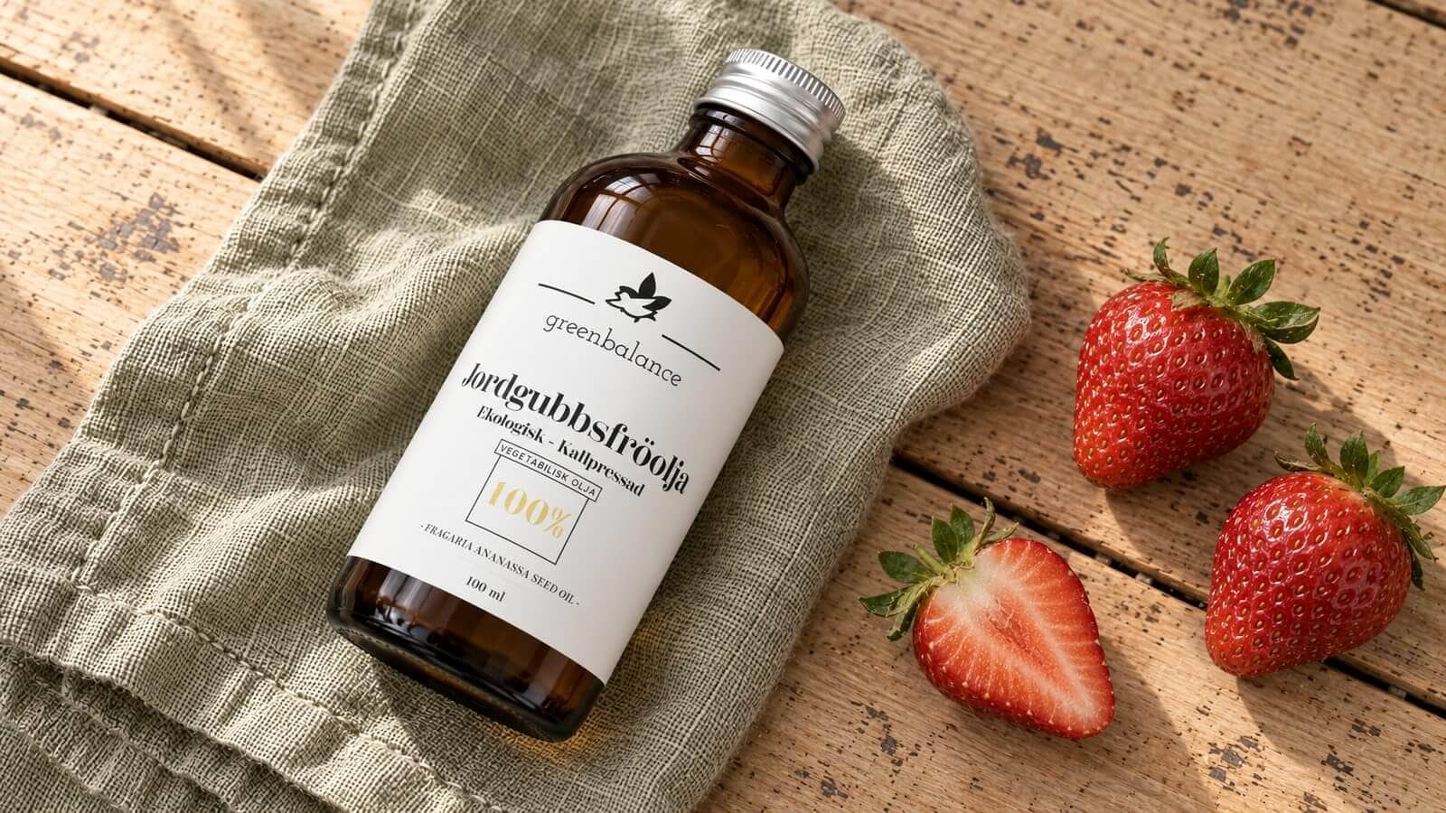 jordgubbsfroolja produkt ravaror Flaska med jordgubbsfröolja liggande på linne och trä, med hela och delade jordgubbar.