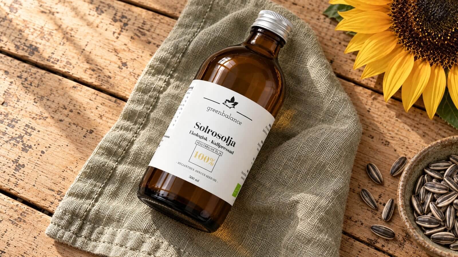 solrosolja produkt ravaror Flaska med solrosolja liggande på linne och trä, med solros och solrosfrön.