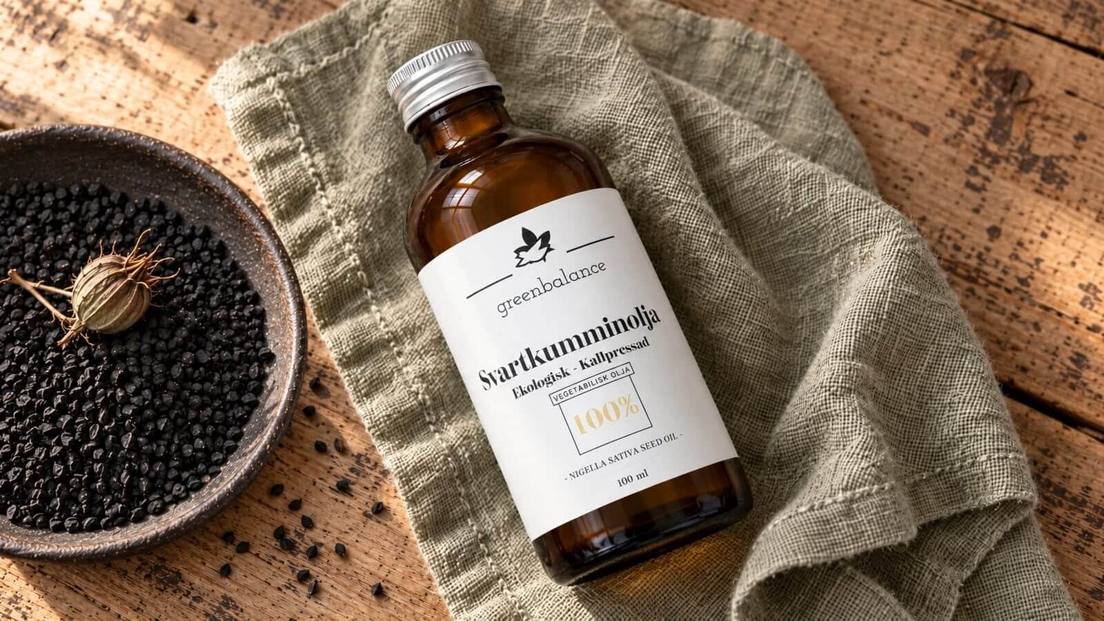 svartkumminolja produkt ravaror Flaska med svartkumminolja på linne och trä intill en bred mörk skål med svarta frön och en torr frökapsel.